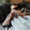 Man laying on couch with blanket on looking at his phone.