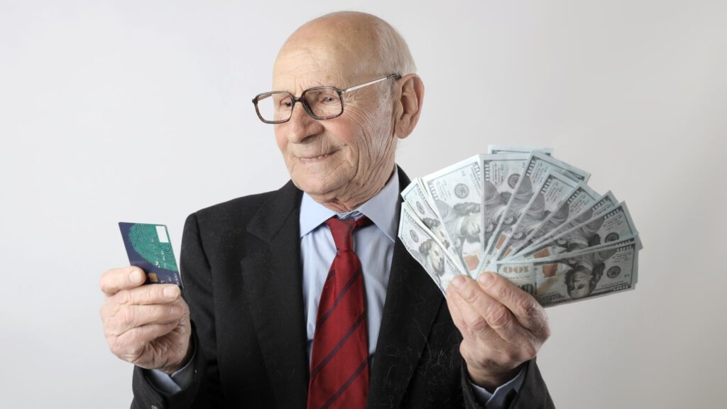 An old man in a suit holding cash and a credit card.