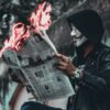 A man in a Guy Faux mask reading a burning newspaper.